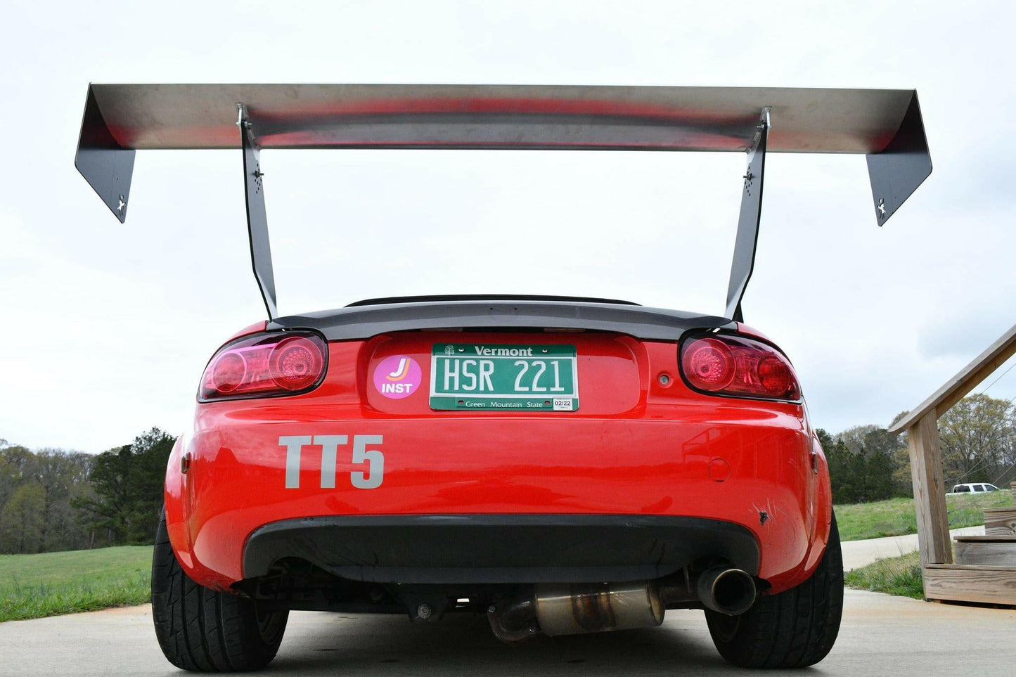 A red sports car with a Vermont license plate that reads "HSR 221" is parked outdoors on a cloudy day, with grass and trees in the background. The car showcases the Miata Big Wang Kit '06-15 NC (V2) from 9livesracing for enhanced racing aerodynamics, dual exhaust pipes, and the label "TT5" on the back.
