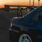 Sunset view of the rear half of a dark-colored sports car featuring a prominent rear spoiler and white wheels. The car, equipped with the 9livesracing Evolution 8/9 Big Wang kit ’03-07 CT9A down-force aluminum wings, is parked on an empty asphalt lot. The blurred horizon and sky in the background showcase hues of orange and pink.