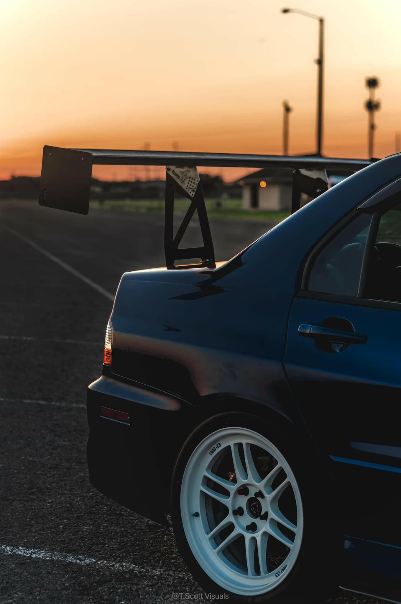 Sunset view of the rear half of a dark-colored sports car featuring a prominent rear spoiler and white wheels. The car, equipped with the 9livesracing Evolution 8/9 Big Wang kit ’03-07 CT9A down-force aluminum wings, is parked on an empty asphalt lot. The blurred horizon and sky in the background showcase hues of orange and pink.