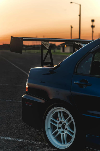 Sunset view of the rear half of a dark-colored sports car featuring a prominent rear spoiler and white wheels. The car, equipped with the 9livesracing Evolution 8/9 Big Wang kit ’03-07 CT9A down-force aluminum wings, is parked on an empty asphalt lot. The blurred horizon and sky in the background showcase hues of orange and pink.