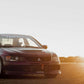 A black Mitsubishi Lancer Evolution with a 9livesracing Evolution 8/9 Big Wang kit '03-07 CT9A aluminum wing is parked on a dirt road at sunset. The sun is low on the horizon, casting a warm glow over the scene. The car has racing decals on the windshield and doors, ensuring optimal down-force for high-speed stability.