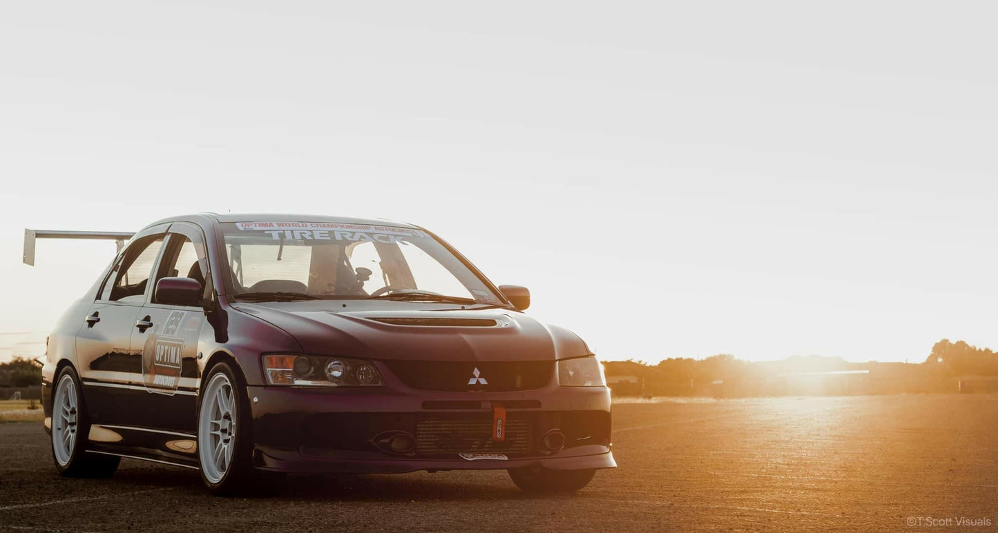 A black Mitsubishi Lancer Evolution with a 9livesracing Evolution 8/9 Big Wang kit '03-07 CT9A aluminum wing is parked on a dirt road at sunset. The sun is low on the horizon, casting a warm glow over the scene. The car has racing decals on the windshield and doors, ensuring optimal down-force for high-speed stability.