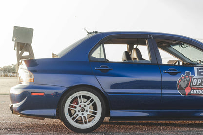 A blue sports car, featuring a 9livesracing Evolution 8/9 Big Wang kit ’03-07 CT9A with a rear wing and partially visible racing decals on the side door, is parked on a road. The car showcases white alloy wheels designed for maximum down-force, set against an open, sunlit landscape.