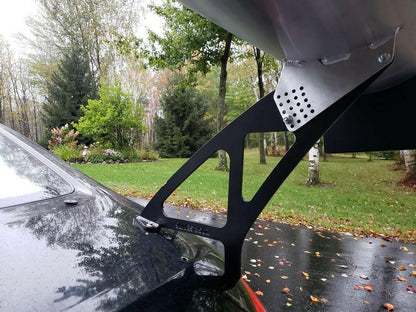Close-up of a 350z Big Wang Kit '02-08 Z33 from 9livesracing mounted on the trunk of a black car, featuring an extruded aluminum wing supported by a metal bracket. The car, showcasing Big Wang GT3 performance, is parked outdoors on a wet driveway, surrounded by lush trees and green shrubs under an overcast sky.