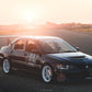 A sleek black Mitsubishi Lancer Evolution, boasting racing decals and a 9livesracing Evolution 8/9 Big Wang kit ’03-07 CT9A rear spoiler, is parked on an empty road at sunset. The car features white wheels, various sponsor stickers including "Tire Rack" and "Optima," and aluminum wings enhancing its down-force as the sun casts a warm glow.