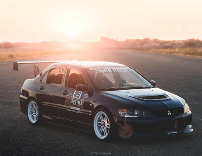 A sleek black Mitsubishi Lancer Evolution, boasting racing decals and a 9livesracing Evolution 8/9 Big Wang kit ’03-07 CT9A rear spoiler, is parked on an empty road at sunset. The car features white wheels, various sponsor stickers including "Tire Rack" and "Optima," and aluminum wings enhancing its down-force as the sun casts a warm glow.