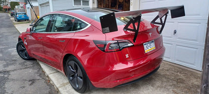 A red Tesla Model 3, equipped with a large aftermarket Big Wang GT3 rear wing/spoiler from 9livesracing, is parked in a residential area. The car features black wheels and a tinted rear window, enhancing its sleek appearance. Residences and garages are visible in the background while the license plate reads "CS5-3417.
