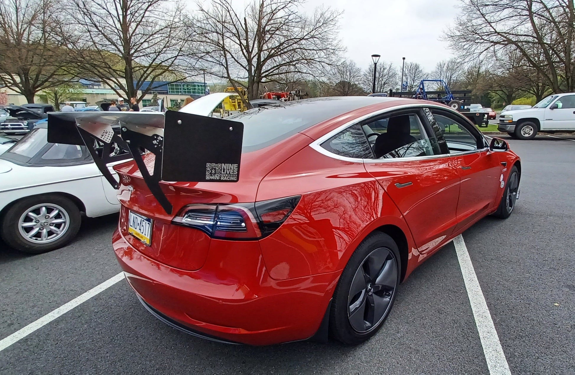A red Tesla Model 3 equipped with a 9livesracing Big Wang kit, featuring a large aftermarket rear wing spoiler, reminiscent of a GT3, is parked in a lot. The background includes other vehicles and leafless trees under a cloudy sky. The car's license plate and the added text on the extruded aluminum wings are visible.