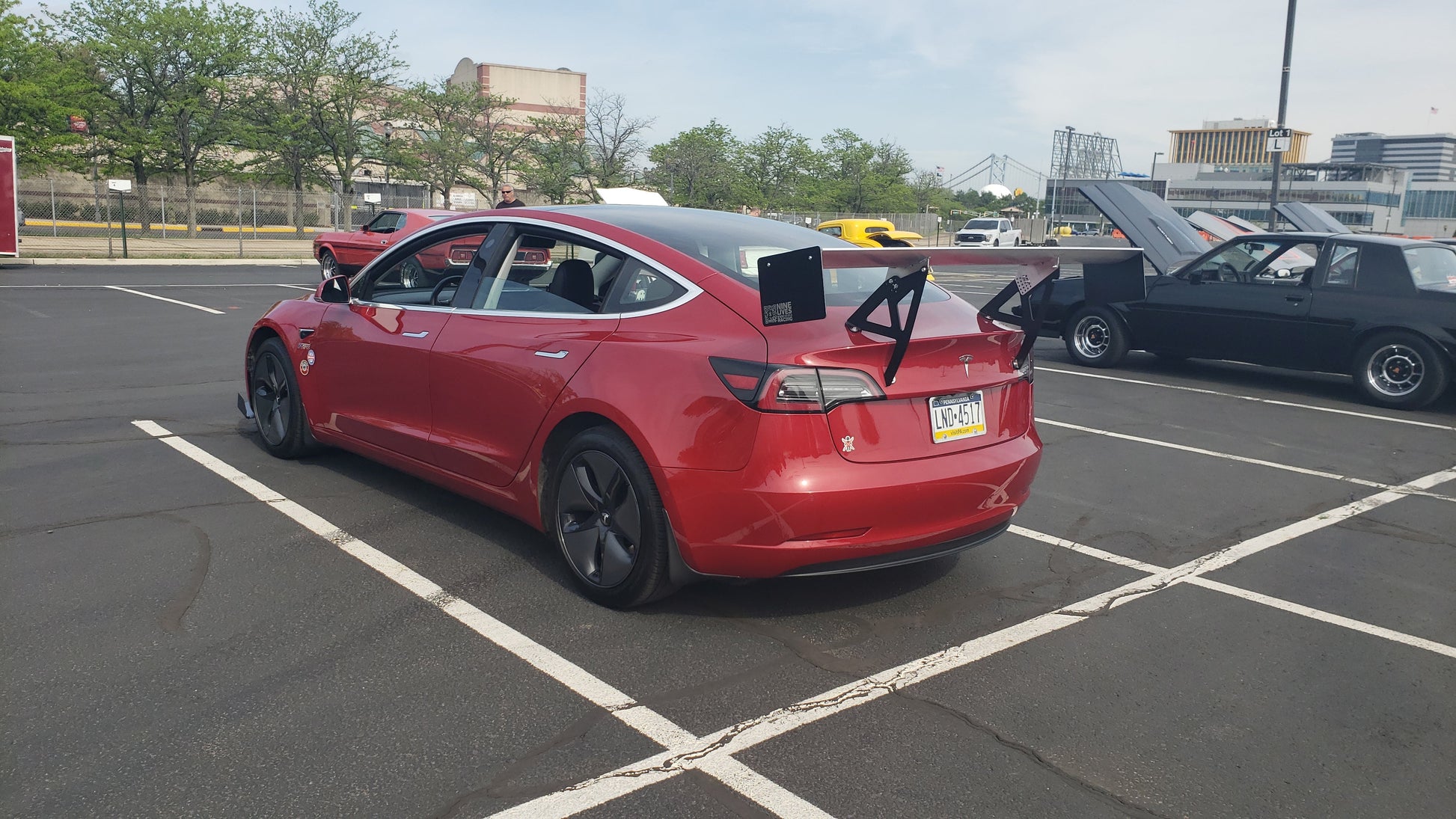 A red Tesla Model 3, equipped with a 9livesracing Big Wang GT3 spoiler made from extruded aluminum and bearing a Pennsylvania license plate, is parked in a lot at a car show. The trunk is open along with the hoods of some other cars, hinting at modifications or displays. Trees and a building are visible in the background.