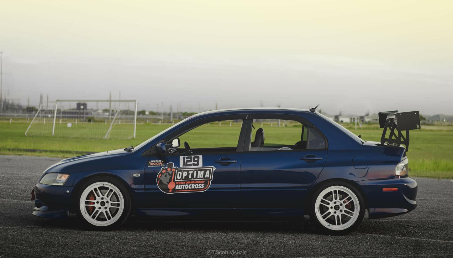 A dark blue sports car equipped with the 9livesracing Evolution 8/9 Big Wang kit ’03-07 CT9A rear wing, white rims, and racing number "129" on the side door parked on a racetrack. The car displays various sponsor decals, including an "Optima Autocross" sticker. In the background, a fenced field and distant landscape can be seen.