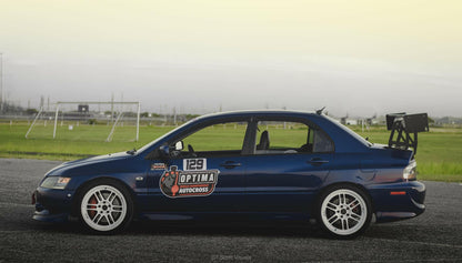 A dark blue sports car equipped with the 9livesracing Evolution 8/9 Big Wang kit ’03-07 CT9A rear wing, white rims, and racing number "129" on the side door parked on a racetrack. The car displays various sponsor decals, including an "Optima Autocross" sticker. In the background, a fenced field and distant landscape can be seen.