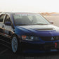 A blue Mitsubishi Lancer Evolution is parked on an open road, showcasing a hood vent, white rims, and various decals, including one indicating participation in the Optima World Championship. In the warm light of sunset, its 9livesracing Evolution 8/9 Big Wang kit '03-07 CT9A aluminum wings stand out, designed for maximum down-force.