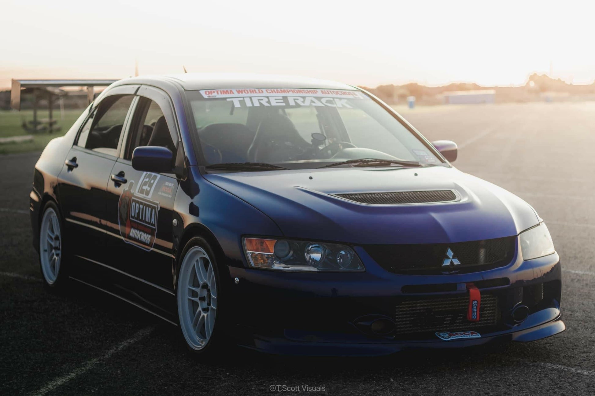 A blue Mitsubishi Lancer Evolution is parked on an open road, showcasing a hood vent, white rims, and various decals, including one indicating participation in the Optima World Championship. In the warm light of sunset, its 9livesracing Evolution 8/9 Big Wang kit '03-07 CT9A aluminum wings stand out, designed for maximum down-force.