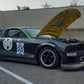 A black race car with the number 89 on its door sits parked with its hood open. The car boasts various sponsor logos and prominently features a Mustang Big Wang Kit '05-09 S197 from 9livesracing as its performance rear wing. It is parked in an outdoor lot with barriers and a metal building in the background.