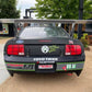 A black Ford Mustang race car adorned with decals such as "TOYO TIRES," "NASA," and "89 AI" is parked outdoors. Equipped with the 9livesracing Mustang Big Wang Kit '05-09 S197 for optimal downforce adjustments, the car also features a message on the trunk reading "Good Food Good Friends Good Times." The backdrop includes trees, a picnic area, and modern buildings.