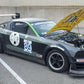 A race car with number 89 displayed on the side is parked in a lot, its hood open to reveal the engine. It boasts features like the Mustang Big Wang Kit '05-09 S197 by 9livesracing for optimal downforce adjustments, along with "American Iron" and various sponsor decals. Nearby, a person is holding a mobile phone.