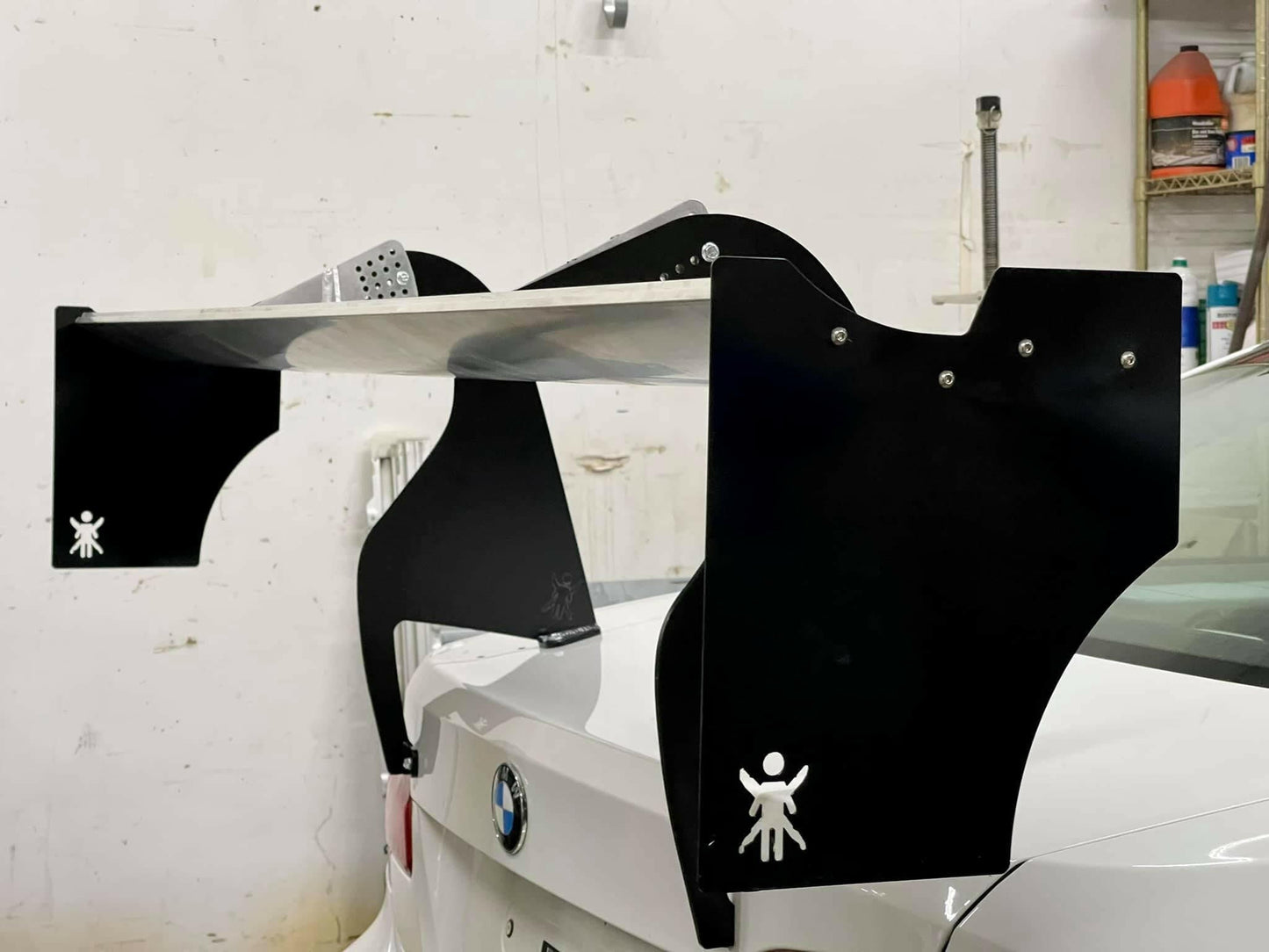 A large spoiler is mounted on the rear of a white BMW car. The spoiler features black End Plates for The Big Wáng! by 9livesracing, with a logo of a stick figure holding up its arms. Engineered for optimal downforce, the aluminum setup complements the garage backdrop filled with tools and equipment.