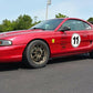A red Mustang Big Wang Kit '94-‘98 SN95 from 9livesracing, adorned with a black number "11" in a white circle decal on the door and several other racing decals, bronze wheels, and extruded aluminum wings, is parked on a paved driveway. In the background, a white vehicle is visible next to a grassy area and trees.