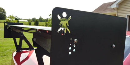 Close-up of a black metal Mustang Big Wang Kit '94-'98 SN95 rear wing from 9livesracing on a red car. The wing features an extruded aluminum design with a cutout of a running person. The background showcases a grassy yard with trees and a beige house with a slanted roof.