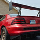 A Mustang equipped with the 9livesracing Big Wang Kit for '94-‘98 SN95, featuring a large rear wing, black rear diffuser, extruded aluminum wings, a number #8 decal on the side, and bronze-colored wheels is parked on a driveway in front of a garage. The rear license plate reads "9848JS," showcasing its aerodynamic design for an optimal lift-to-drag ratio.