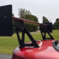 A close-up of a Mustang Big Wang Kit '94-'98 SN95 by 9livesracing, featuring extruded aluminum wings attached to the trunk of a red sports car. The background showcases a lawn, trees, and a paved driveway under a cloudy sky.