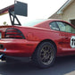 A red race car with a large rear spoiler and the number 77 on the door is parked in front of a garage. The car features various decals and modifications, including racing wheels, USA flag, blue tow hooks, and an impressive Mustang Big Wang Kit '94-‘98 SN95 by 9livesracing for optimal lift to drag ratio. Another vehicle and a beige house are visible in the background.