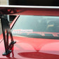 A close-up of the rear section of a red car, showcasing a Mustang Big Wang Kit '94-‘98 SN95 rear wing spoiler mounted on the trunk. The spoiler features extruded aluminum wings and is supported by black brackets, enhancing its lift-to-drag ratio. A "9livesracing" decal is visible on the back window. In the background, part of a garage and yard can be seen.