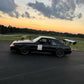 A Mustang Big Wang Kit '79-‘93 Fox body by 9livesracing, featuring a sleek black finish and a prominent rear wing for optimal down-force along with the racing number “717” on the door, is parked on a paved surface. The background showcases a grassy field and a picturesque sunset with scattered clouds in the sky.