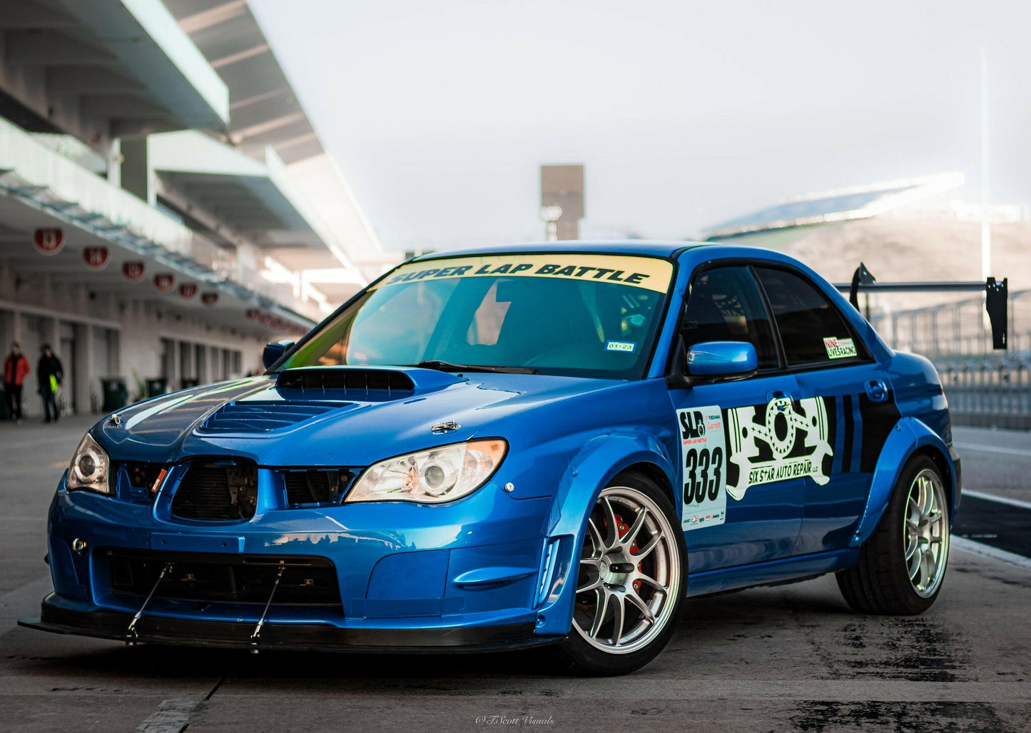 A blue Subaru Impreza STI Big Wang kit '00-07 GD from 9livesracing, featuring a black front splitter and a large aluminum rear wing, is parked on the track. It boasts numerous racing decals, including "Super Lap Battle" on the windshield and "333" on the side. The background reveals a bustling racing circuit and grandstands, emphasizing its high-octane downforce design.