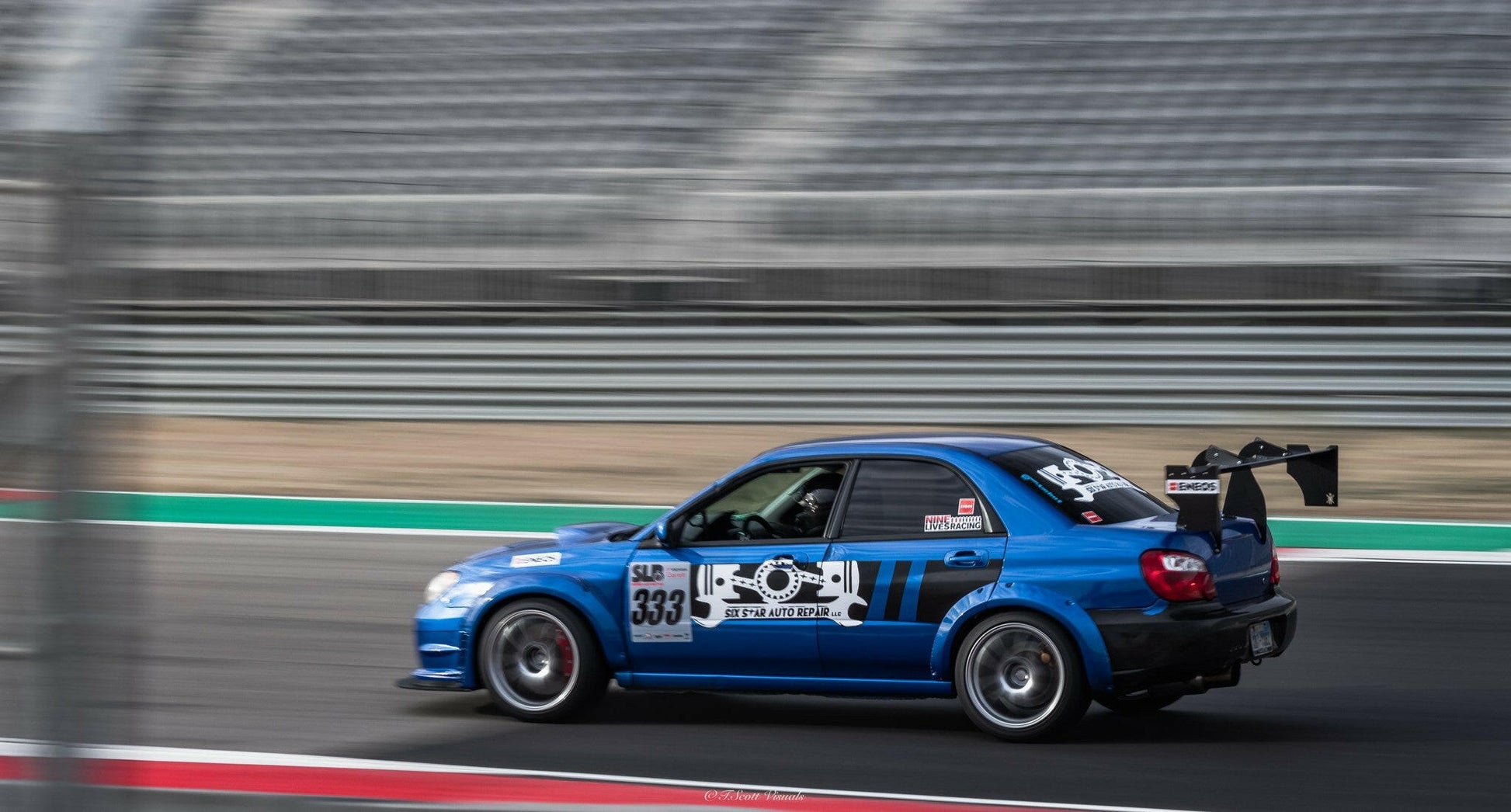 A blue Subaru Impreza STI featuring the 9livesracing Big Wang kit ('00-'07 GD) with the number 335 on its doors zooms down the track, sporting multiple sponsor decals and a substantial airfoil spoiler for maximum downforce. The image beautifully captures the car's motion with a blurred effect, set against empty grandstands in the background.