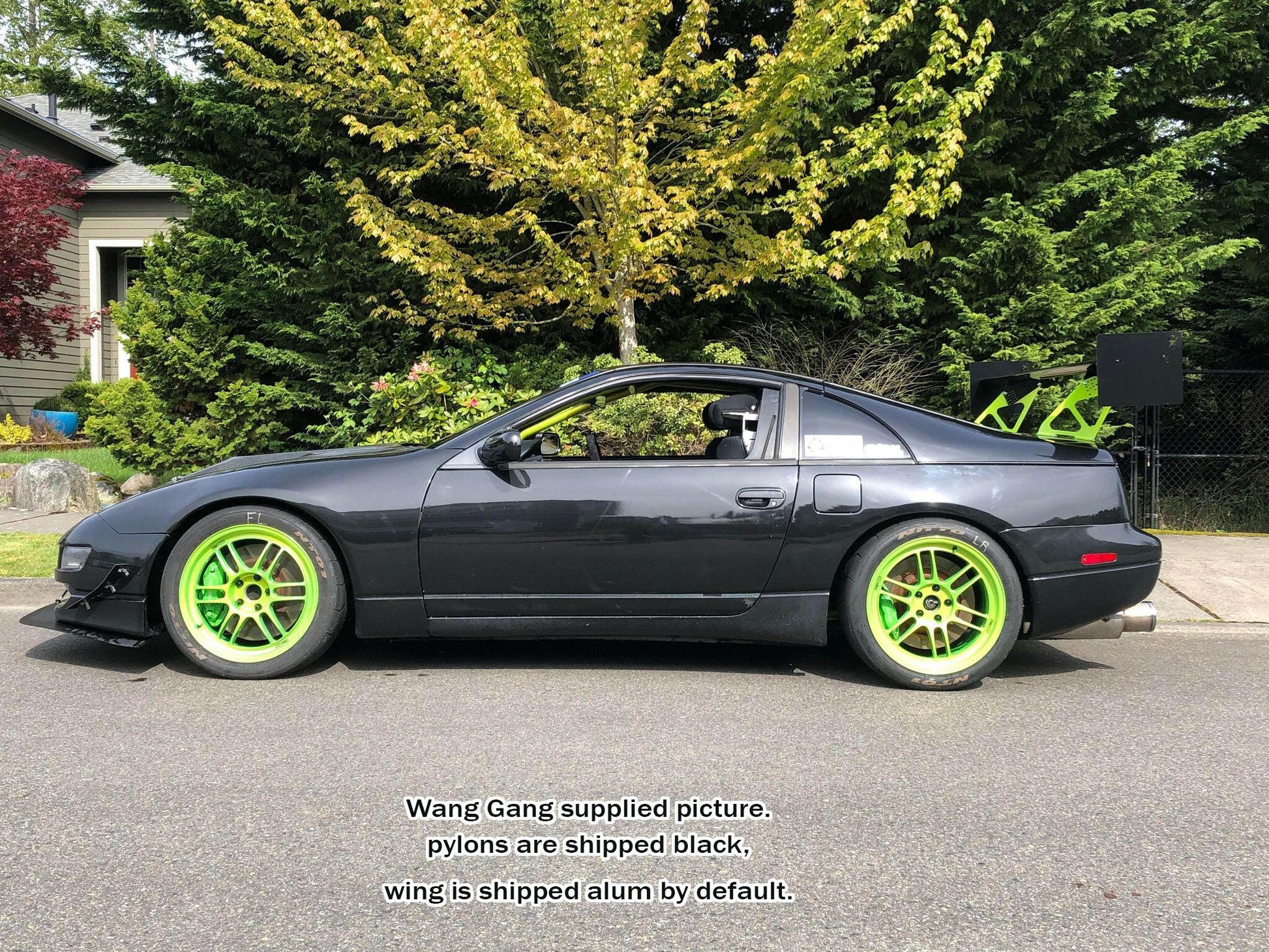 A slick black sports car with lime green rims and a 9livesracing 300ZX Big Wang kit '88-00 Z32 rear wing, captured on a residential street. Text over the image reads: "Wang Gang supplied picture. Pylons are shipped black, Big Wang GT3 wing is shipped alum by default." The background features trees and bushes.