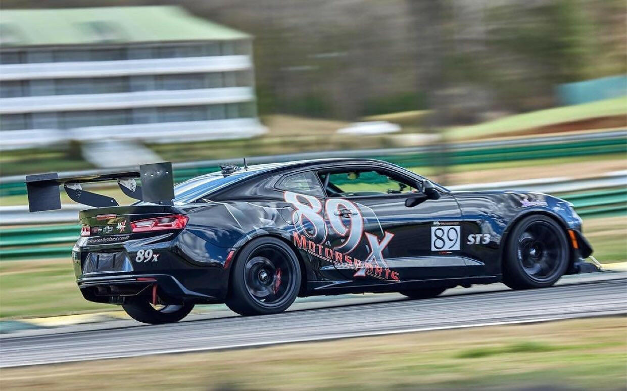 The Camaro Wang 2016+ 6th Gen from 9livesracing, bearing the number 89 and "80x Motor Sports" on the sides, speeds down a race track. The black race car features aluminum wings for enhanced downforce and sleek black wheels. In the background, a building and greenery indicate an outdoor racing event, showcasing its impressive GT3 performance.