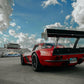 A low-angle shot of a red race car featuring "The Deuce! Carbon Double Element Kit" by 9livesracing, parked on a track. The car has a "RUSH2R" license plate. The sky above is partly cloudy, with bleachers and other vehicles visible in the background.