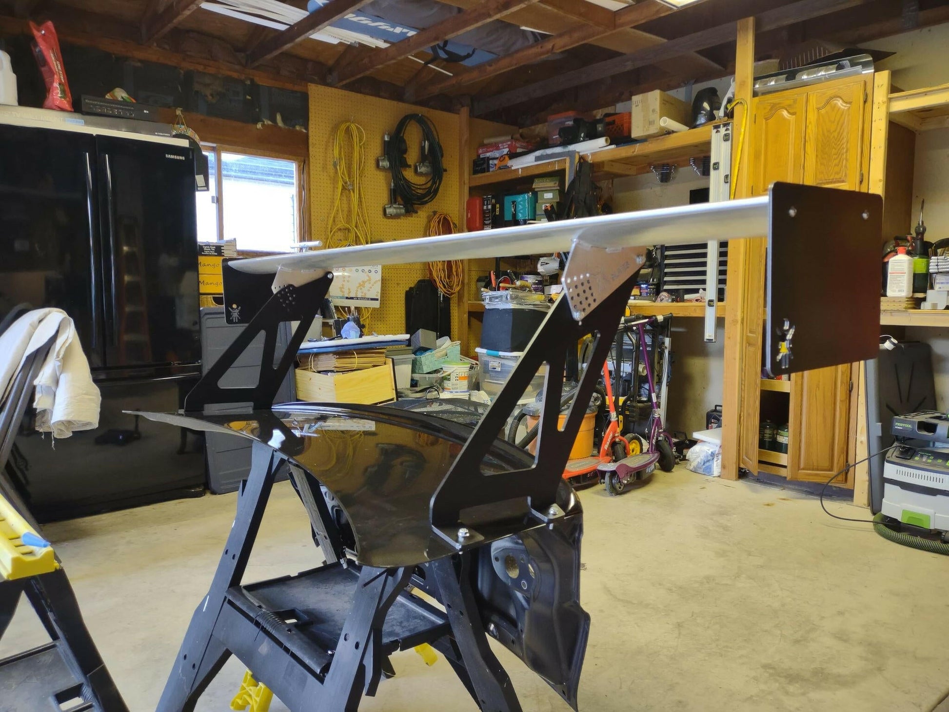 The Evolution 10 Big Wang kit, by 9livesracing, features a large aluminum wing that generates significant down-force and is mounted on a black trunk lid in a cluttered garage. Shelves and tools are visible in the background, along with a black refrigerator and wooden cabinets. The garage appears to be a workspace for car modifications.
