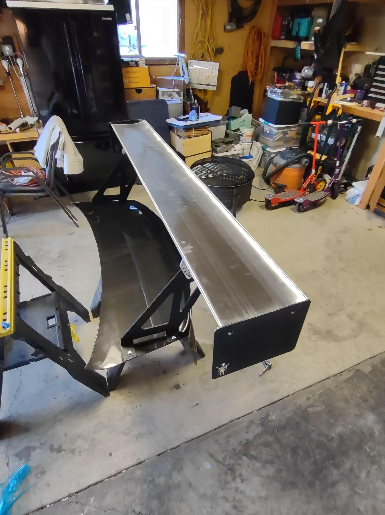 An Evolution 10 Big Wang kit by 9livesracing, likely crafted from aluminum to enhance down-force for an automobile, is installed indoors, possibly in a garage or workshop. The wing is supported by a black frame amidst scattered tools and equipment; nearby are a lawnmower, shelves stocked with various supplies, and extension cords hanging on the wall.
