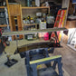 A cluttered garage with tools, shelves, and various items. In the center, a large black Evolution 10 Big Wang kit by 9livesracing is being worked on, placed on a sawhorse with a power drill nearby. A red ladder leans against the back wall, and various storage boxes are visible on the shelves.
