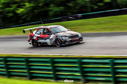 An Is300 Big Wang Kit 98-04 xe10 by 9livesracing, featuring a sleek black and red race car with number 100 on the door and equipped with a Big Wang GT3 spoiler for optimal down-force adjustments, speeds around a track with green barriers, surrounded by lush greenery. The vivid camouflage design stands out as blurred lines in the background indicate high speed.