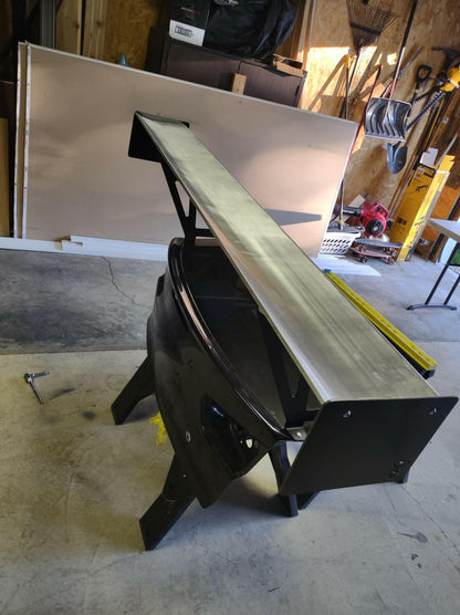 In a garage or workshop, the Evolution 10 Big Wang kit from 9livesracing, a large black metal spoiler nicknamed the Big Wang, stands upright on a concrete floor. Behind it, an assortment of tools, wooden panels, and construction materials are visible. A wrench placed near the spoiler suggests recent adjustments for achieving optimal down-force.