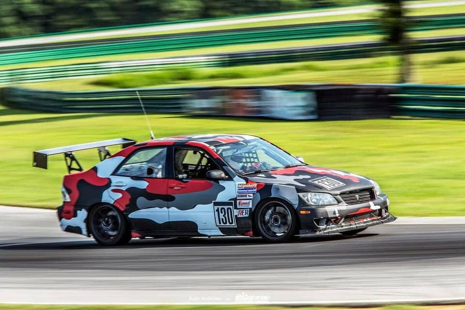 A race car with a striking red, black, gray, and white camouflage pattern speeds around a racetrack. Equipped with the Is300 Big Wang Kit 98-04 xe10 from 9livesracing for optimal down-force adjustments, it proudly displays the number 130 on its side. Various sponsor decals adorn its exterior. Green barriers and blurry foliage in the background suggest high speed.