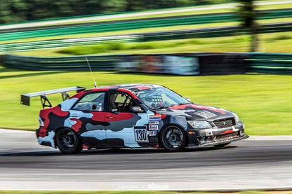 A race car with a striking red, black, gray, and white camouflage pattern speeds around a racetrack. Equipped with the Is300 Big Wang Kit 98-04 xe10 from 9livesracing for optimal down-force adjustments, it proudly displays the number 130 on its side. Various sponsor decals adorn its exterior. Green barriers and blurry foliage in the background suggest high speed.