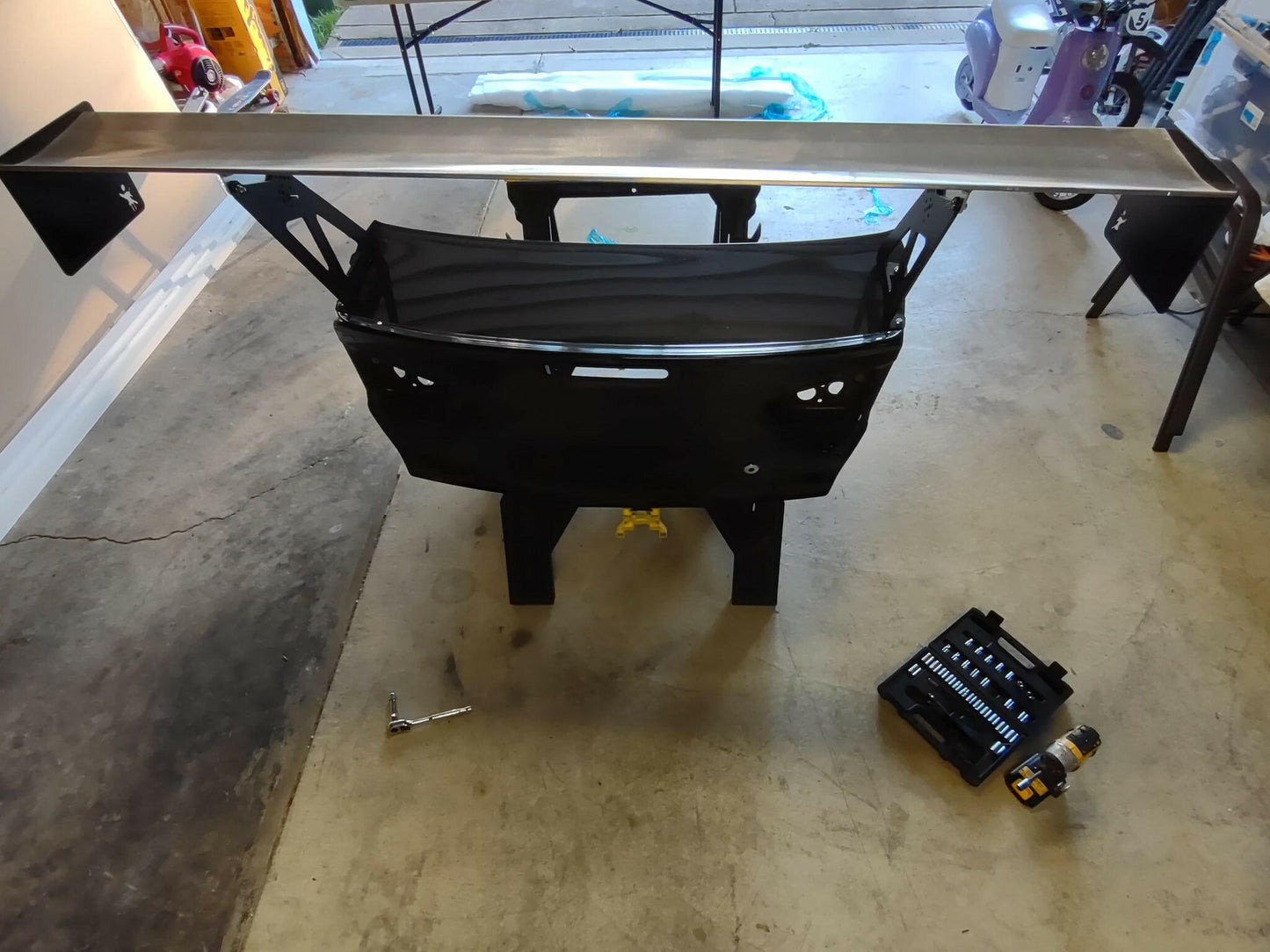 A workbench with an Evolution 10 Big Wang kit from 9livesracing being assembled. Tools and equipment, including a wrench, a toolkit, and a power drill, are placed on the concrete floor around the workbench. The setting appears to be a garage with various items in the background, highlighting an impressive aluminum wing designed for maximizing down-force.