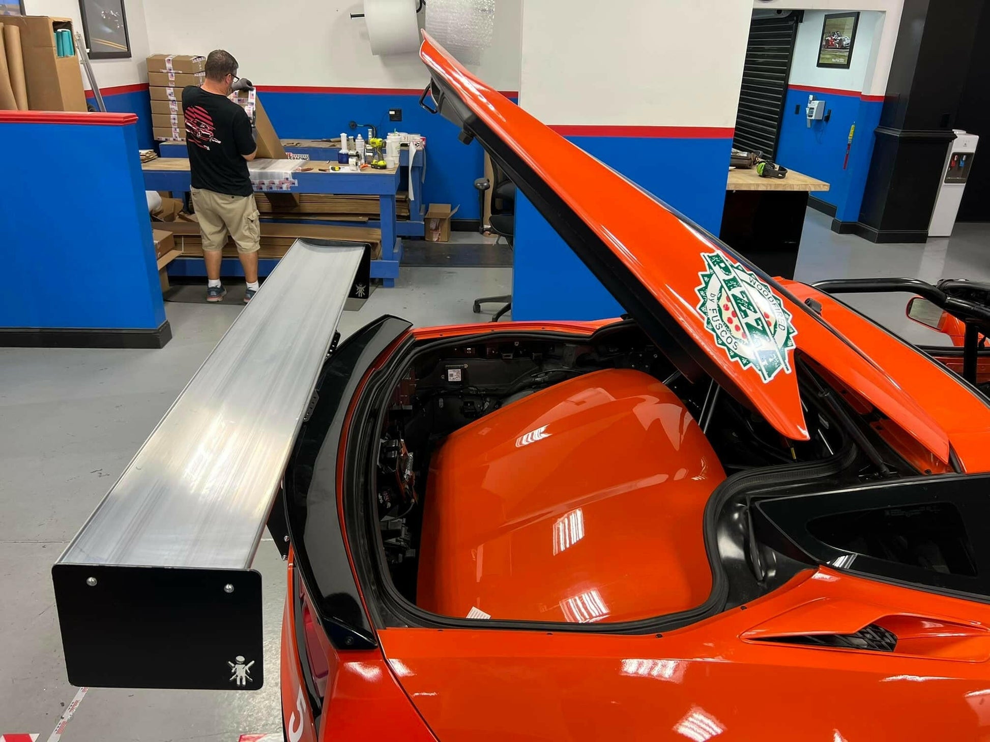 A vibrant orange sports car with an open rear engine compartment, revealing its components. A large silver spoiler, resembling the 9livesracing Corvette Big Wang Chassis Mount for 2014 - 2019 C7 models, is attached to the back. In the background, a person in a workshop setting is making down-force adjustments next to a wooden table filled with supplies.