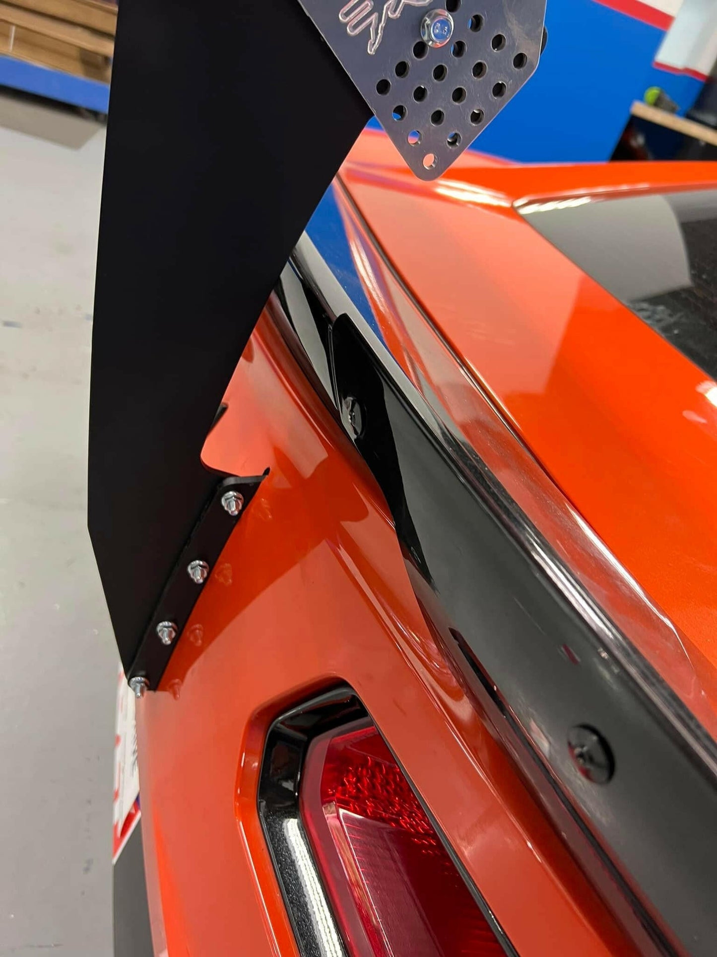Close-up of a Corvette Big Wang Chassis Mount ’2014 - 2019 C7 by 9livesracing, featuring down-force adjustments and aluminum wings attached to the trunk. The glossy orange vehicle showcases its metallic bracket and black sidepieces, with part of a red and white car light also visible.
