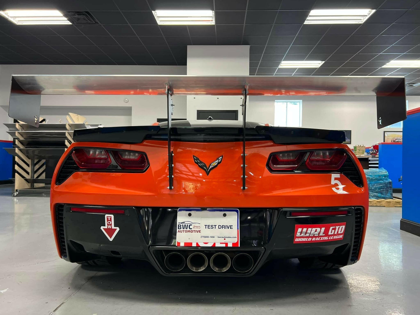 A vibrant orange Corvette Big Wang Chassis Mount '2014 - 2019 C7 by 9livesracing with a large GT3 rear wing and quad exhaust pipes is parked inside a garage. The rear license plate reads "TEST DRIVE." The car has racing decals, including a prominent "5" and "WWR GTO" label. Various workshop tools and equipment are visible in the background.