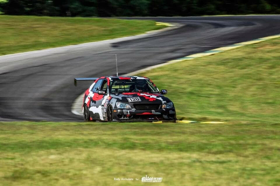 A race car sporting a black, red, gray, and white camouflage paint scheme zips down a curving track. Outfitted with the Is300 Big Wang Kit 98-04 xe10 from 9livesracing for optimal downforce adjustments and a front splitter, it handles the turns effortlessly. The backdrop features lush green grass and trees lining the course.