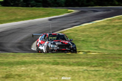 A race car sporting a black, red, gray, and white camouflage paint scheme zips down a curving track. Outfitted with the Is300 Big Wang Kit 98-04 xe10 from 9livesracing for optimal downforce adjustments and a front splitter, it handles the turns effortlessly. The backdrop features lush green grass and trees lining the course.