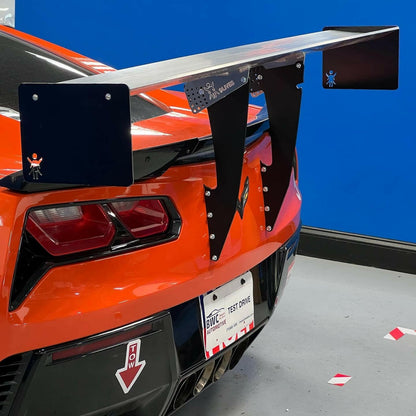 An orange sports car, outfitted with the remarkable 9livesracing Corvette Big Wang Chassis Mount designed for the 2014 - 2019 C7 model, is shown from the rear. The car is indoors against a blue wall, featuring a "Test Drive" label on its license plate. Small decals of a waving stick figure adorn the aluminum wings of the impressive spoiler.