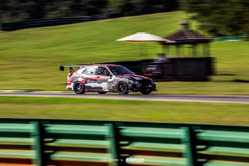An Is300 with a camouflage design speeds down the track, motion blur accentuating the speed. The background features a green, grassy field, a gazebo structure, and another car slightly behind. Green and yellow barriers line the track in the foreground as the 9livesracing Is300 Big Wang Kit 98-04 xe10 benefits from optimal down-force adjustments.