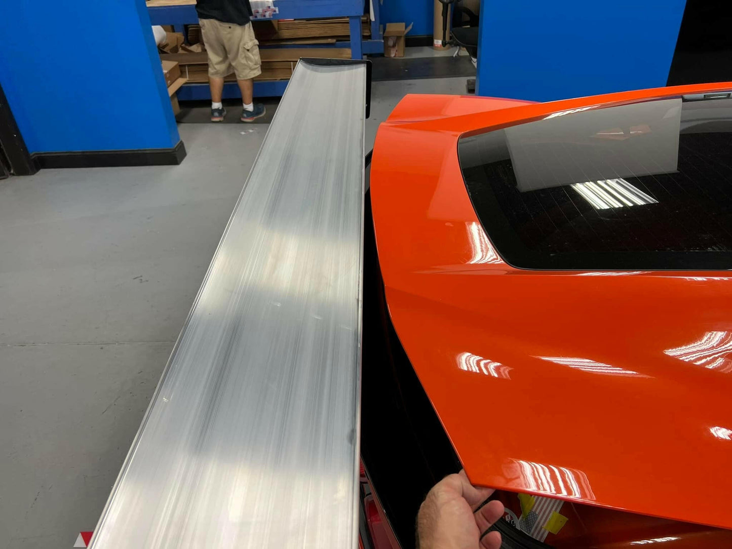 Close-up of an orange Corvette sports car equipped with a 9livesracing Big Wang Chassis Mount for ’2014 - 2019 C7 models, parked in a garage. In the background, a person wearing shorts and sneakers stands near shelves stocked with various items. The workshop or garage space features blue walls and a grey floor.