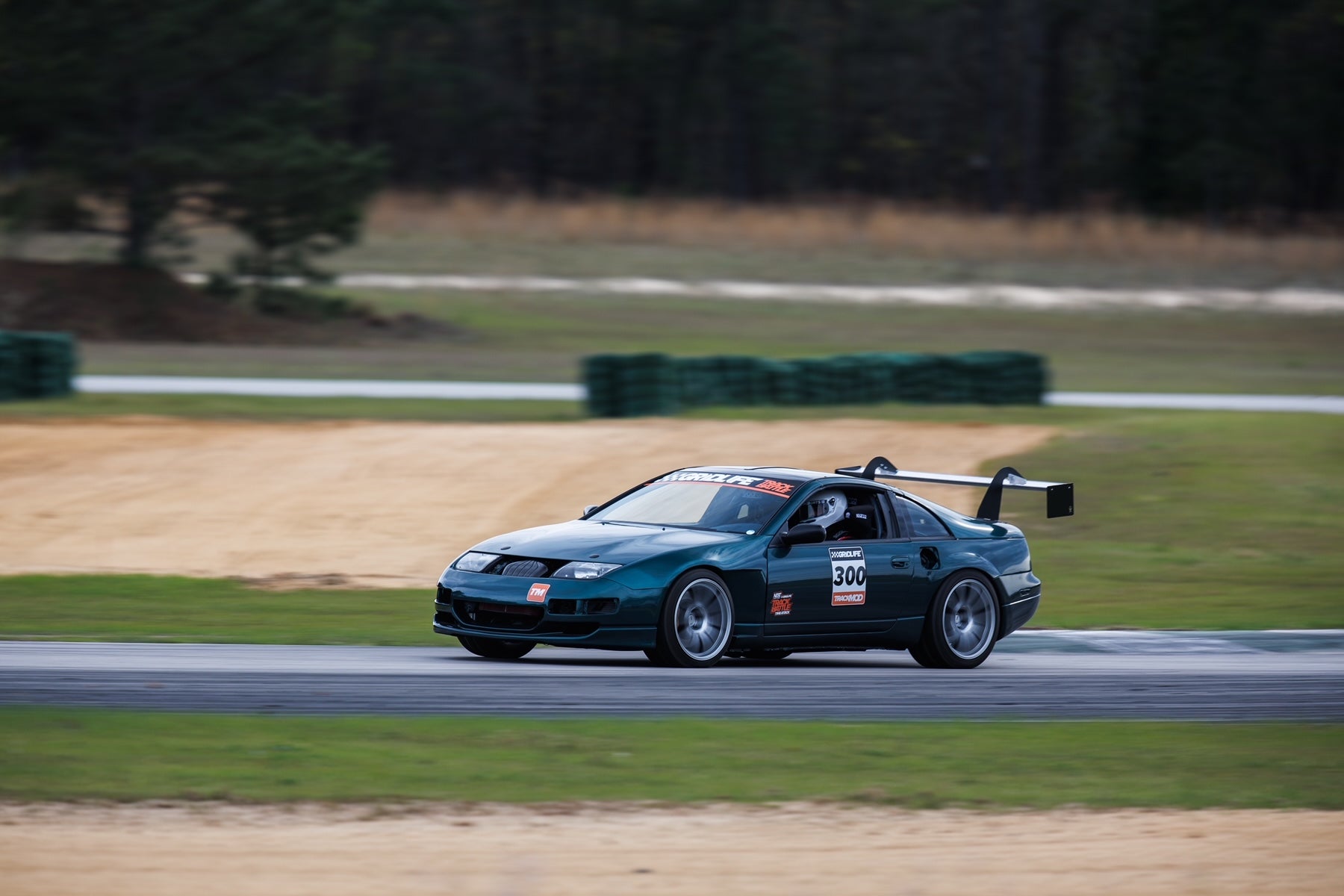 A dark green sports car, adorned with racing decals and the number 300, speeds along a racetrack. The car, equipped with the 300ZX Big Wang kit '88-00 Z32 from 9livesracing for optimal down-force, is captured making a turn with a blurred background, indicating motion. The surroundings include grassy areas and safety barriers.